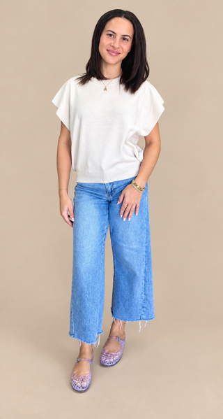 A woman modeling a white short-sleeve top and wide-leg blue jeans against a solid beige studio background.