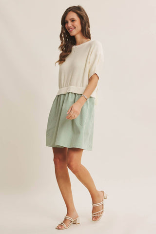 A woman modeling a short-sleeve cream knit top and a light sage green pleated mini skirt, paired with beige strappy heeled sandals, against a plain light beige studio background.