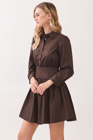 A woman modeling a dark brown long-sleeve mini dress with a button-down collar, gathered waist, and flared skirt against a plain light background.