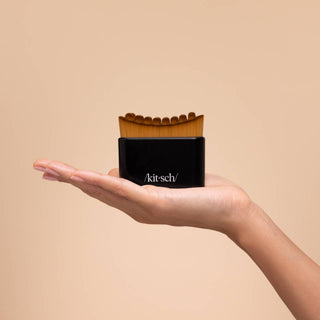 A hand holding a small black cosmetic container with a wavy wooden applicator, branded /kit-sch/, against a solid beige background.
