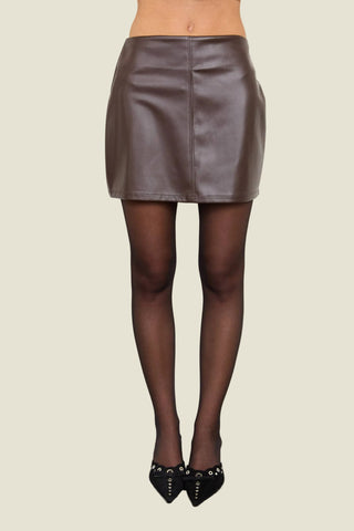 A model's lower body wearing a brown faux-leather mini skirt, sheer black tights, and black studded heels, standing against a plain off-white wall.