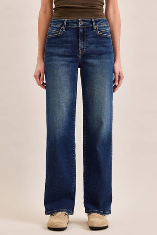 A person wearing dark blue wide-leg jeans, a brown ribbed top, and beige shoes, standing against a plain beige studio background.