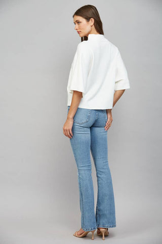 A woman shown from the back wearing a white short-sleeve blouse and light blue flare jeans against a plain gray studio background.