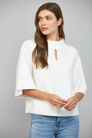 A woman wearing a white mock-neck top with wide flared sleeves and a small keyhole cutout at the collar, paired with blue jeans, against a solid gray studio background.