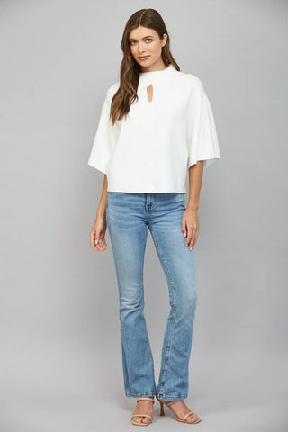 A woman modeling a white short-sleeve top with a keyhole cutout and light-wash blue flare jeans against a solid gray studio background.