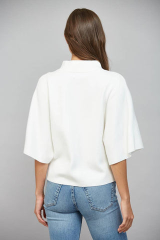 A woman viewed from the back wearing a white short-sleeve knit top with a high mock neck and blue jeans, set against a solid gray studio background.