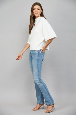A woman modeling a white short-sleeve blouse with a high mock neck and keyhole cutout, paired with light-wash blue jeans and nude heeled sandals, against a solid gray studio background.