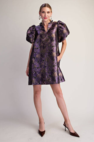 A woman modeling a short, knee-length purple dress with a metallic gold floral jacquard pattern, puffed short sleeves, and a high collar, paired with dark brown pointed-toe heels against a light gray studio background.