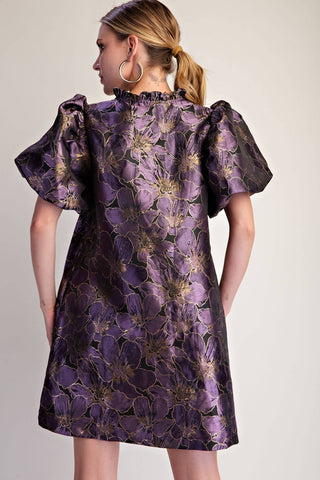 A woman shown from the back wearing a short-sleeved purple dress with a metallic gold floral jacquard pattern, photographed against a neutral light beige background.