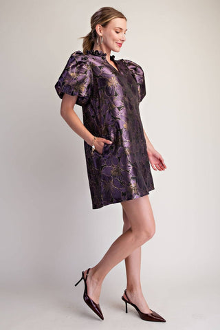 A woman models a short, puffed-sleeve mini dress with a purple and gold floral jacquard pattern, paired with dark brown pointed-toe slingback heels against a light gray studio background.