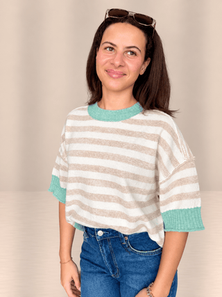 A woman wearing a beige and white horizontally striped knit top with mint green ribbed collar, cuffs, and hem, paired with blue jeans and sunglasses on her head, set against a plain beige background.