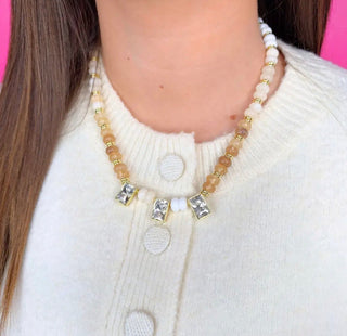 A close-up of a person wearing a beaded necklace with gold-toned rectangular bezel settings and clear stones, paired with a cream knit cardigan. The necklace features alternating translucent peach-beige and white beads with small gold spacer beads.