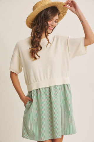 A woman wearing a cream short-sleeve knit top and a light green and white vertically striped skirt, styled with a straw hat against a warm beige studio background.