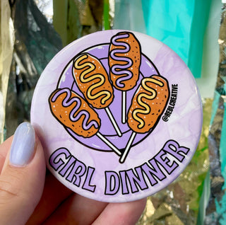 Button badge with corn dogs and 'Girl Dinner' text held by a hand.