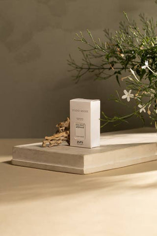 A minimalist white rectangular product box labeled 'HONEY MOIST' and 'puro' sits on a light stone pedestal, styled with dried branches and green flowering stems against a warm beige background. Studio Megee Sea Salt Driftwood fragrancei refill bottle and packaging on a beige background