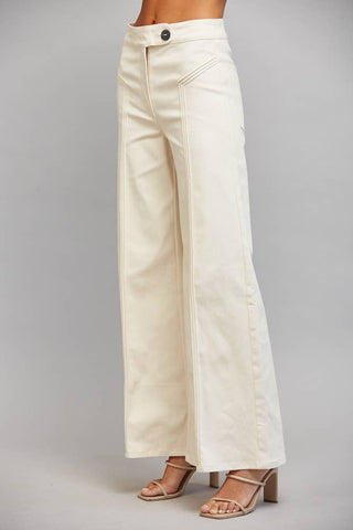 A model is shown from the waist down wearing off-white wide-leg pants and beige strappy high-heeled sandals against a neutral gray studio background.