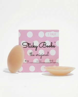 A pink polka-dot box labeled 'Sticky Boobs the original' with two flesh-toned adhesive nipple covers placed in front on a clean white background.