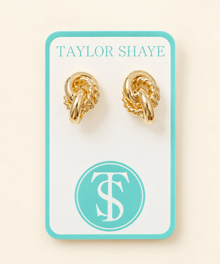 A pair of gold knot-shaped hoop earrings displayed on a white jewelry card with a turquoise border. The card is branded 'TAYLOR SHAYE' at the top and features a circular 'TS' logo at the bottom, set against a light cream background.