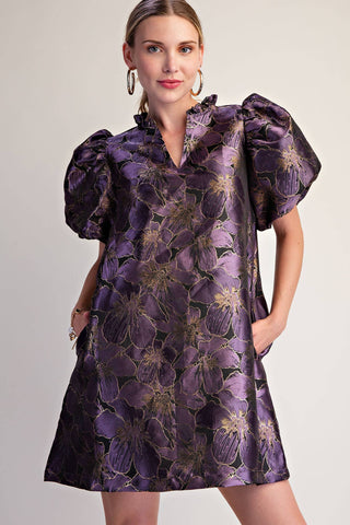A woman modeling a short purple satin dress with a large floral pattern and voluminous puff sleeves, set against a plain light gray studio background.