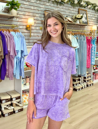 A woman modeling a matching lavender-purple tie-dye lounge set consisting of a short-sleeve top with a chest pocket and matching shorts, standing inside a retail clothing store.