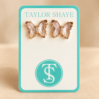 A pair of butterfly-shaped stud earrings with pink faceted crystals and gold-tone settings, displayed on a white card with a turquoise border. The card is branded 'TAYLOR SHAYE' at the top and features a matching circular 'TS' logo at the bottom, set against a neutral beige background.