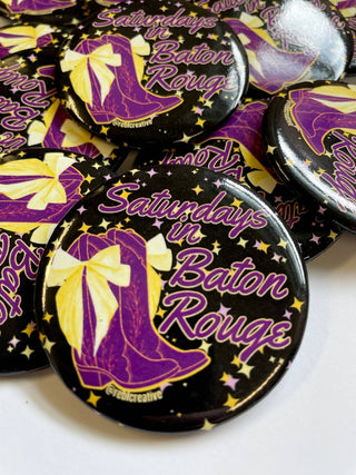 A pile of round pin-back buttons featuring a purple cowboy boot with a yellow bow on a black background, surrounded by yellow stars and the text 'Saturdays in Baton Rouge' in purple script.