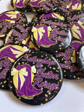 A pile of round pin-back buttons featuring a black background with purple and yellow graphics. The design shows a purple boot with a yellow bow, gold stars, and the text 'Saturns in Baton Rouge' in purple script.