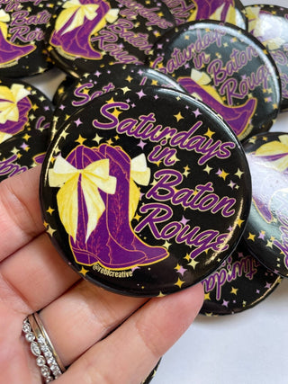 A close-up of a hand holding a round pin-back button badge with the text 'Saturdays in Baton Rouge' in purple script over a black background with gold stars and a purple-and-gold boot illustration. Other matching badges are scattered in the background.