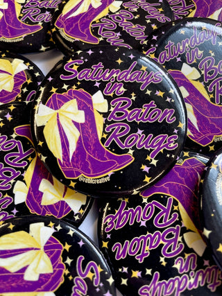 A close-up pile of round pinback buttons featuring the phrase "Saturdays in Baton Rouge" in purple script over a black background with gold stars and a purple-and-gold high-heel illustration.