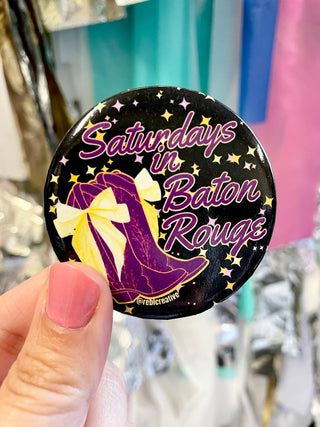 A hand with pink nail polish holds a round pin badge featuring a purple high-heel boot with a yellow bow on a black starry background, with the text 'Saturdays in Baton Rouge' in purple script.