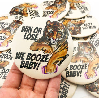 Button badge with tiger illustration and 'We Booze Baby!' text held in a hand, surrounded by more badges.
