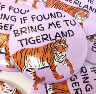 A close-up of round pin-back buttons featuring a purple background with the phrase 'IF FOUND, BRING ME TO TIGERLAND' in black text and an orange-and-white tiger illustration.