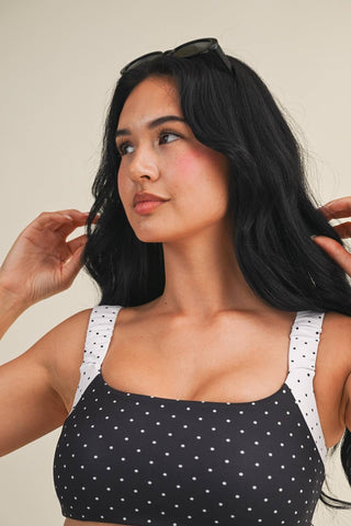 Woman wearing a black polka dot top with white straps against a neutral background