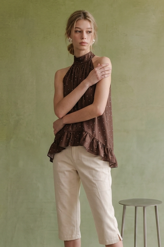 A woman modeling a sleeveless high-neck brown top with small white polka dots and a ruffled hem, paired with light beige cropped pants, standing against a textured sage green wall.