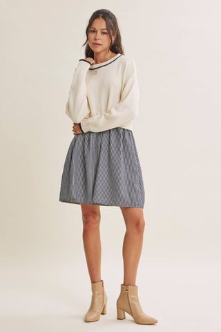 A woman modeling a cream knit sweater with a navy and white striped pleated mini skirt and beige ankle boots, standing against a solid light beige studio background.