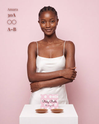 A woman stands against a pink background wearing a white camisole dress, with two small pink and white bra cups displayed on a white pedestal in front of her. Text on the left reads 'Amara 30A A-B' and the cup packaging is visible on the pedestal.