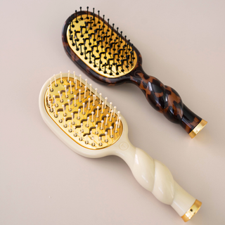 Two hairbrushes on a light beige background. One has a tortoiseshell pattern handle with black and brown tones, and the other has a cream-colored handle with gold accents.