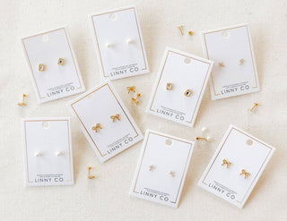 Gold earrings on LINNY CO branded cards on a beige background