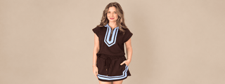A woman modeling a dark brown short-sleeve tunic dress with light blue and white geometric trim details on the chest and hem, set against a solid beige studio background.