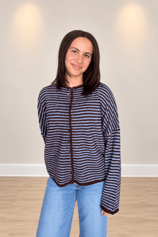 A woman wearing a navy and dark brown horizontally striped zip-up cardigan with dark brown trim, paired with light blue jeans, standing in a room with a beige wall and light wood floor.