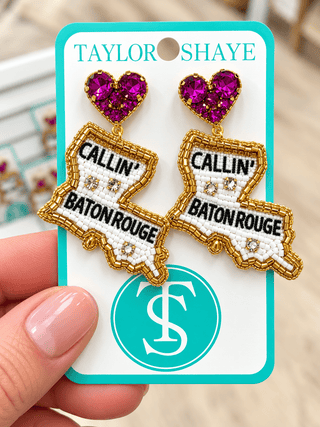 Taylor Shaye embroidered patch earrings shaped like the state of Louisiana, featuring a bright pink heart-shaped rhinestone stud and white, black, and gold embroidered details with 'CALLIN' BATON ROUGE' text, shown on a branded display card held by a hand.