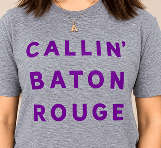 A close-up of a person wearing a heather gray t-shirt with the phrase 'CALLIN' BATON ROUGE' printed in bold purple letters across the front.