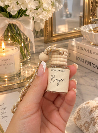 A hand holds a small glass reed diffuser bottle labeled 'HOLLOWWOOD Boujee' in a warm, styled interior setting with a lit candle, flowers, books, and a mirror in the background.