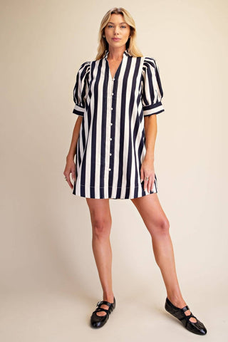 A woman modeling a short black and white vertically striped button-up shirt dress with puff sleeves, paired with black Mary Jane flats, against a solid beige studio background.