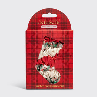 A retail package of Kit Sches Ruched Satin Scrunchieles in a Christmas stocking design, featuring a red and black plaid box with a clear window showing the cream satin scrunchies with red bows and green holly accents.