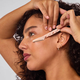 Woman having her eyebrows shaped with a razor by another person.