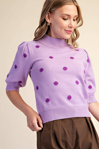A woman wearing a lavender purple knit mock neck sweater with darker purple textured polka dots, paired with brown trousers, against a solid beige background.