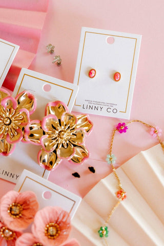 A styled flat-lay of jewelry on a pink background, including pink and gold flower-shaped earrings, matching stud earrings on white cards labeled LINNY CO, a gold chain necklace with colorful beads, and pink paper flowers.