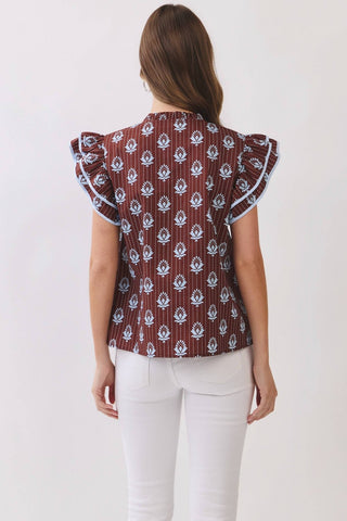 A woman viewed from the back wearing a brown ruffled blouse with a white floral block print and white pants, set against a plain off-white background.