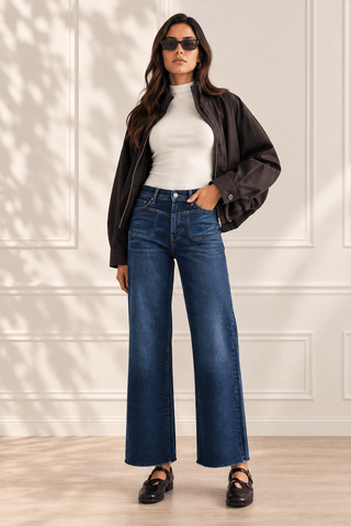 A woman modeling a pair of high-waisted, wide-leg blue jeans with a white mock neck top, a black bomber jacket, and black Mary Jane shoes, standing against a white paneled wall.